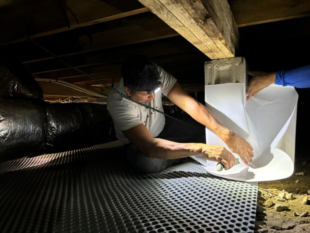 Hawk Crawlspace & Foundation Repair technician installing crawl space encapsulation material in a dimly lit crawl space, highlighting moisture control and home protection in Chesapeake, VA.